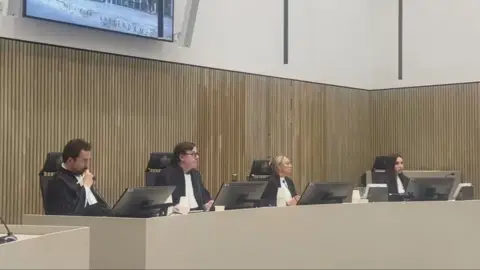 A courtroom with three judges sitting, plus a furth person taking information down.