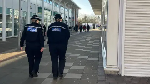 Two female police officers walking down a street in Telford. The photograph is taken from the back so you can't see their faces. They're both wearing black police uniforms and police bowler hats. The officer on the left has a blue and white label reading 'POLICE' on the back of her coat, whilst the person on the right has one that reads 'POLICE COMMUNITY SUPPORT OFFICER.' 