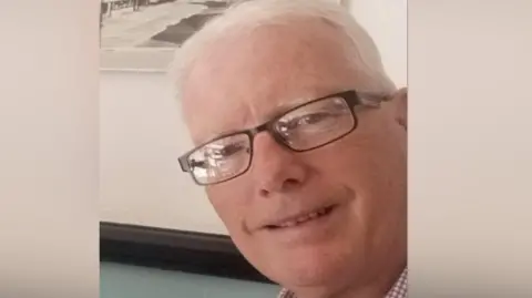 Andrew Avery smiling for the camera. He is wearing glasses. There is a picture on the wall behind him.