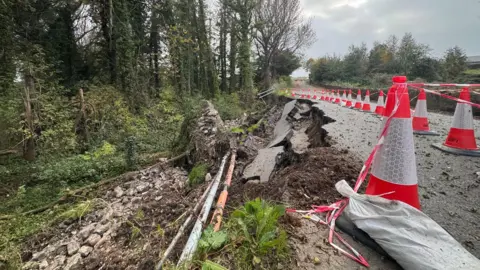 A wide shot of the Maydown Road bridge - in the foreground and to the right of the shot are a row of traffic cones cordoning off the collapsed section. To the left is the collapse - the concrete road surface has given way as has an old stone wall. Dirt, rocky debris and exposed cables can be seen. To the left of that is a row of trees.