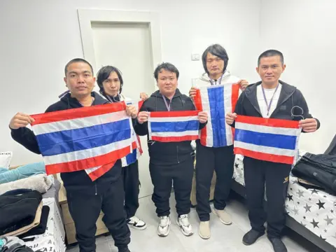 Embassy of the Royal Thai in Tel Aviv The five released hostages stand holding Thai flags in a white room. They are wearing tracksuits and trainers, with lanyards round their necks