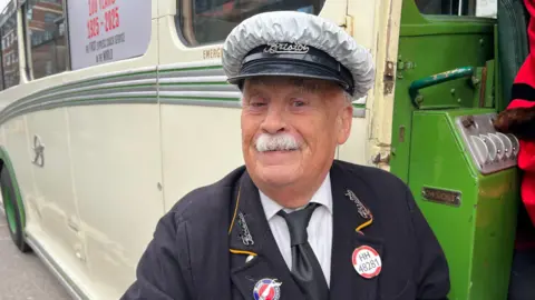 An elderly man with a white moustache looks at the camera. He is wearing a 1920s-era bus drivers uniform and hat. The hat has the word Bristol on it. Part of an historic, cream-coloured coach is visible in the back ground. 