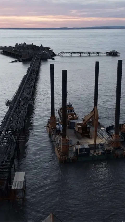 A large specialist barge with four support poles in each corner sits in the water next to Birnbeck Pier at sunset