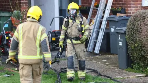 Island Echo Two firefighters wearing breathing apparatus holding fire hoses in the garden of lats with ladders and several bins seen in the garden area.