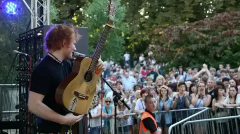 Jenny O'Neill Ed Sheeran performing at Ipswich Music Day in 2009 or 2010. The photo is taken by someone behind or to the side of him on stage. He is standing on stage holding his acoustic guitar. A large crowd can be seen watching on.