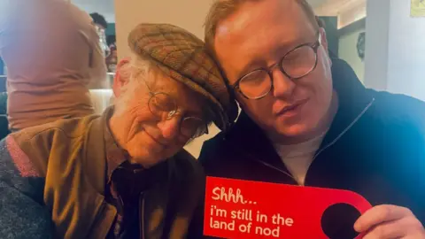 Noddy Holder in a tweed flat cap, glasses and a jacket has his eyes closed as he leans his head towards Tom Seals who also has his eyes closed and is wearing a black quarter zip jumper. He is holding a red sign that reads: Shhh... I'm still in the land of nod