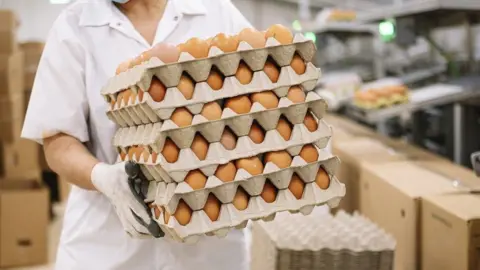 Getty Images Person in a white work uniform and gloves carrying stacked cardboard trays filled with brown eggs inside a food production or packing facility, with boxes and shelves visible in the background.”