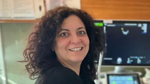 Mid and South Essex NHS Foundation Trust Jennifer Baisden smiles at the camera over her shoulder. She has dark brown curly hair down to her shoulders. A screen can be seen behind her displaying some medical diagrams. 