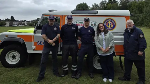 Nicola Parker A large 4-4 that is painted white, grey and orange is parked on grass. In front of it is five people who are smiling at the camera. There are four men and one woman. The men are wearing all navy outfits with a logo on the chest. The woman has dark hair and is wearing a pale green jacket and white t-shirt. 