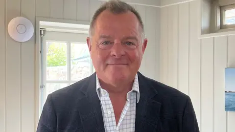 Michael Barnes has short dark hair and is wearing a dark jacket and white checked shirt. He is standing indoors and is looking straight at the camera, smiling.