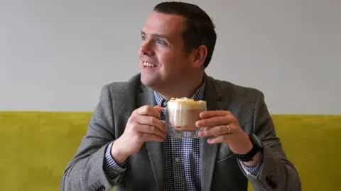PA Media Douglas Ross, a brown-haired man in a grey jacket and blue and white checked shirt, drinks a coffee from a glass cup at a cafe with green velvet seating.