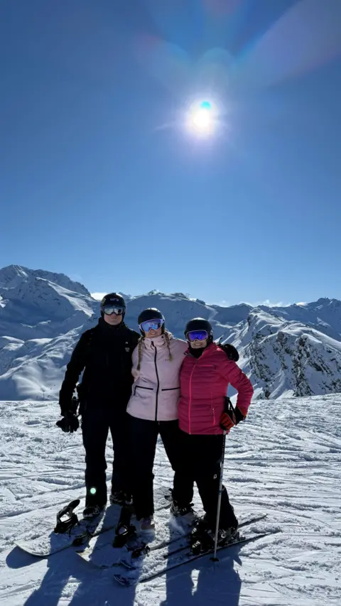Katie Tinkler The image shows three people standing together on a snowy mountain landscape under a clear, bright blue sky. All three individuals are wearing winter sports gear, including helmets, goggles with reflective lenses, insulated jackets, and snow trousers, and each is standing on skis or a snowboard. The person on the left stands on a snowboard, while the two individuals on the right are positioned on skis, with one of them holding a ski pole. Behind the group, there are snow-covered mountain peaks and ridges stretching across the background, with the sunlight shining strongly from above and slightly behind them. The bright sun creates sharp shadows on the snow and highlights the crisp texture of the slopes, giving the scene a vivid, high-contrast appearance with clear visibility and expansive scenery.