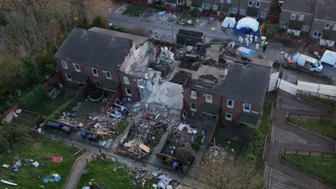 The rear of the houses. The middle of the row shows an exposed concrete interior with rubble outside and debris in the gardens.