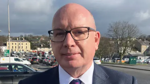 Mr Savage is bald and is wearing rectangular glasses, he has a white shirt and is wearing a black suit with a patterned tie. Behind him is a street where cars are parked along the side and queued up at a set of traffic lights. In the background there are buildings.