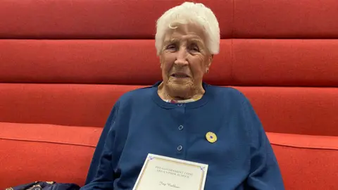Joy Trew smiles at the camera wearing her gold and blue Bletchley medal and holding a note from the Prime Minister's office