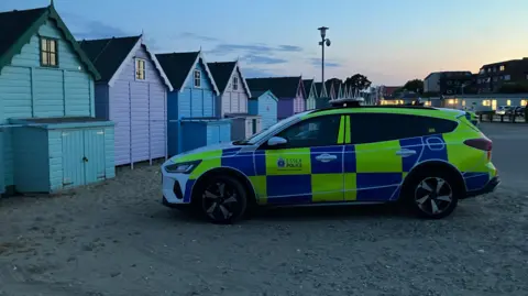 Lewis Adams/BBC An Essex Police estate car parked on a beach. There are colourful beach huts to its left and behind it are buildings, whose lights are on. 