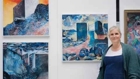 Irina Mackie Artist Tara Harris stands next to paintings displayed on a gallery wall.