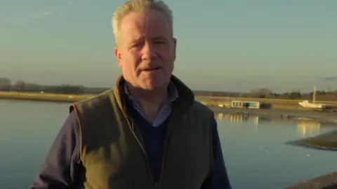 Chris Weston is wearing a green gilet over a blue shirt and jumper and is standing in front of a reservoir.