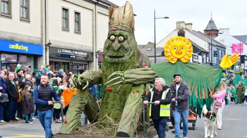 A large tree sculpture covered in moss and red flowers is pulled through Downpatrick town centre. Behind that, is a large sun float with large, outstretched hands. The streets are lined with crowds of people who are watching the parade take place.