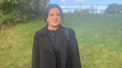 A woman in a black top and a black coat is stood in front of a grass embankment.