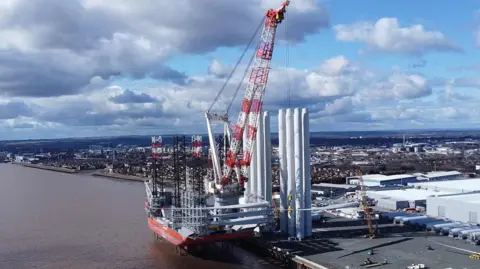 An aerial image of the Wind Peak, a large ship, docked at the Port of Hull. Three large wind turbine blades have been loaded onto the red-coloured vessel which is next to several large white towers. The Humber estuary is visible on the left of the image and the city of Hull is visible in the background.