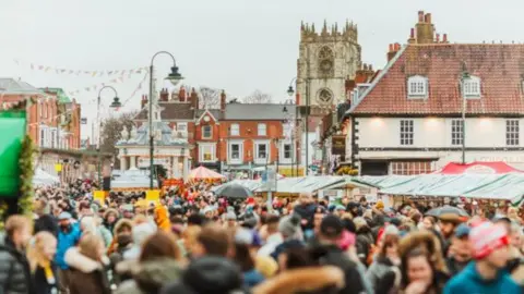 Crowds of people dressed in winter clothing, including some wearing hats, walk past a range of stalls, with a church and other historic buildings in the background.
