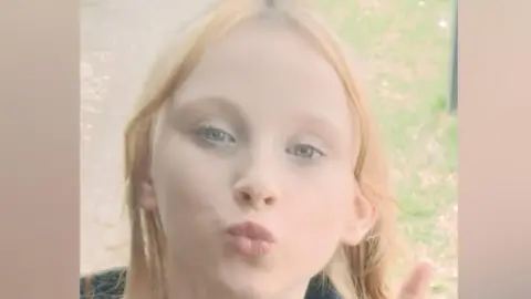 A young girl with light blonde hair is standing outdoors, making a peace sign with her left hand. 
