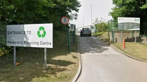 The entrance to Bidston Household Waste Recycling Centre with a large hite sign that says entrance in black writing. A car is travelling along the access road. There is a red circle road sign with 5 written on in black.