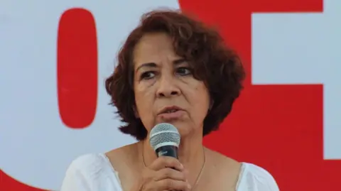 Lilia Gema García Soto holds a microphone as she speaks at a political event. She is wearing a white blouse and behind her, a red-and-white banner can be seen. 