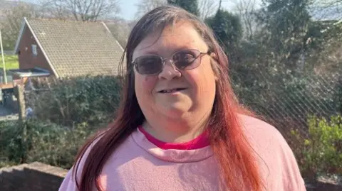 BBC Sam Brown, a woman in her forties with long brown hair dyed red at the ends and wearing a pink top. She is wearing glasses. Behind her is a metal fence with a hedge behind it. The sloping roof of a house can be seen in the left hand side of the shot.