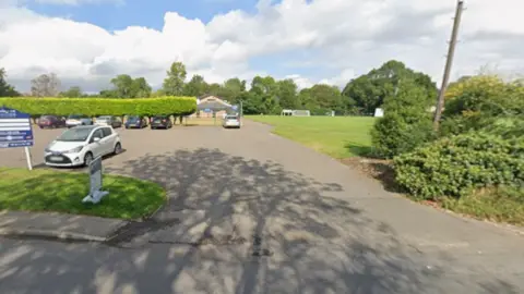Google Cars are parked on a parking area with hedges, a pavilion and playing fields in the background.