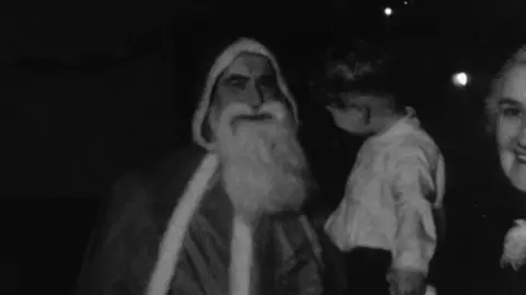Yorkshire Film Archive An old black and white photo of a man dressed as Santa with a hooded cap and a long white beard. He is holding a young boy wearing a shirt and trousers in his arms. Half of a woman's face can be seen on the right of the photo. 