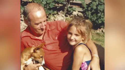 Louise Edwards A man in his forties is lying on the ground and hugging a young girl. She is under one arm and a small dog is under the other arm. It looks like a sunny day and the girl appears to be wearing a swimsuit. The man is wearing a red short sleeved shirt.