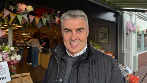 Grace McGrory / BBC Andy Rafters he has short grey hair and is wearing a black padded jacket and grey sweater and white shirt. He stands in front of his greengrocers and flowers shop
