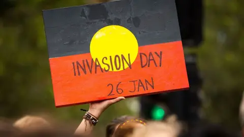 A protestor holds a sign with the red, black and yellow colours of the Aboriginal flag and the words Invasion Day.