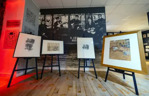 Four pieces of framed artwork are on display on easels in a museum in front of a poster of The Beatles
