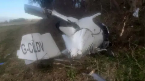 Wreckage of white gyroplane on grass beside field and trees