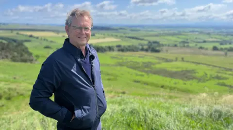 A man with glasses and light brown hair, wearing a blue jacket, looks at the camera. Behind him are green fields