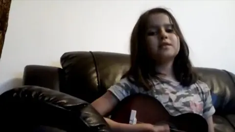 A young girl playing a guitar sits on a black sofa