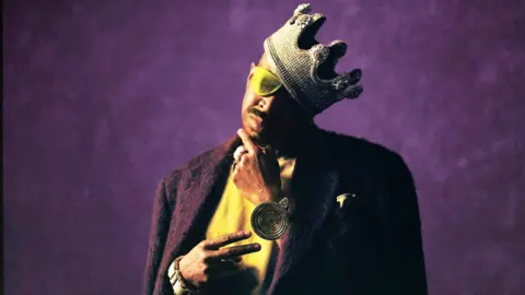 Slick Rick wears a crown and a golden eyepatch, while standing against a regal purple background, in a promotional photo for his 2025 album, Victory