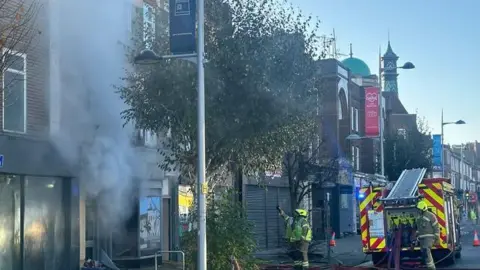Fire crews and an engine on a street in Clacton-on-Sea. Smoke is coming from a building and fire officers are looking and pointing at the building, which is in street full of shops.