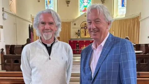 B4SH Two elderly men with long white hair stood in the aisle at a church. They are smiling at the camera