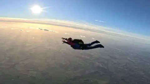 Kayleigh Garbett Kaye mid flight during a lone skydive. She is wearing a helmet, black trousers and a pink sports jacket with a black parachute rucksack on her back. The sun is shining and you can just see the curvature of the earth. There are a few clouds beneath her and all the green fields are miles below on the ground. 