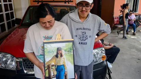 Relatives of América Sánchez hold up a framed photograph of the young cadet in her hometown of Xalapa. in the photo, América Sánchez, is posing in a yellow blouse and jeans in front of a lake. Her relatives are crying. 