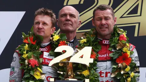 Getty Images Allan McNish on the left of the podium with his two teammates after winning Le Man. They are decked in flowers and are holding a trophy with the number 24 on it.