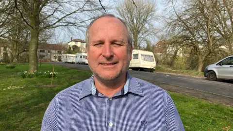 Mike Rigby wearing a blue and white checked shirt looking at the camera in front of some caravans parked on Roman Way in Glastonbury.