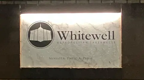 A white sign on a wall which says 'Whitewell Metropolitan Tabernancle'. There is a brown and white symbol of the church as well.