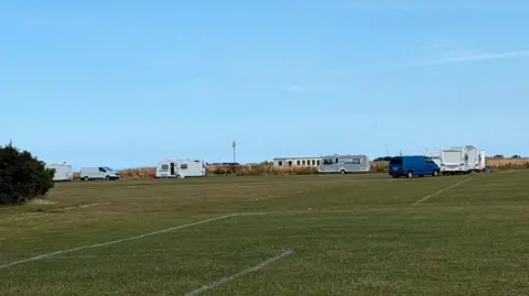 UGC A number of white caravans have been parked around the corner of a football pitch. The caravans are spaced apart with vans between them. A one-storey white building can be seen behind them with a flag mast to the left.