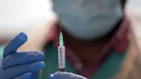 A medical professional in green scrubs and blue gloves preparing a syringe and vaccination to be administered. They are wearing a mask and their face is slightly blurred for effect.