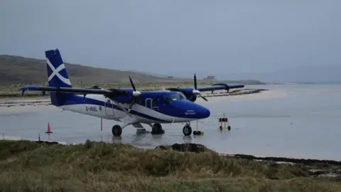 BBC The aircraft is blue and white in colour. It has two propeller engines and is stationary on the sand of Barra's beach airport. It is a grey and chilly day.